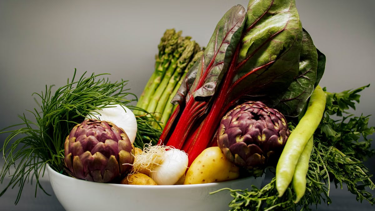 A vibrant assortment of fresh vegetables including artichokes, chard, and asparagus in a white bowl.